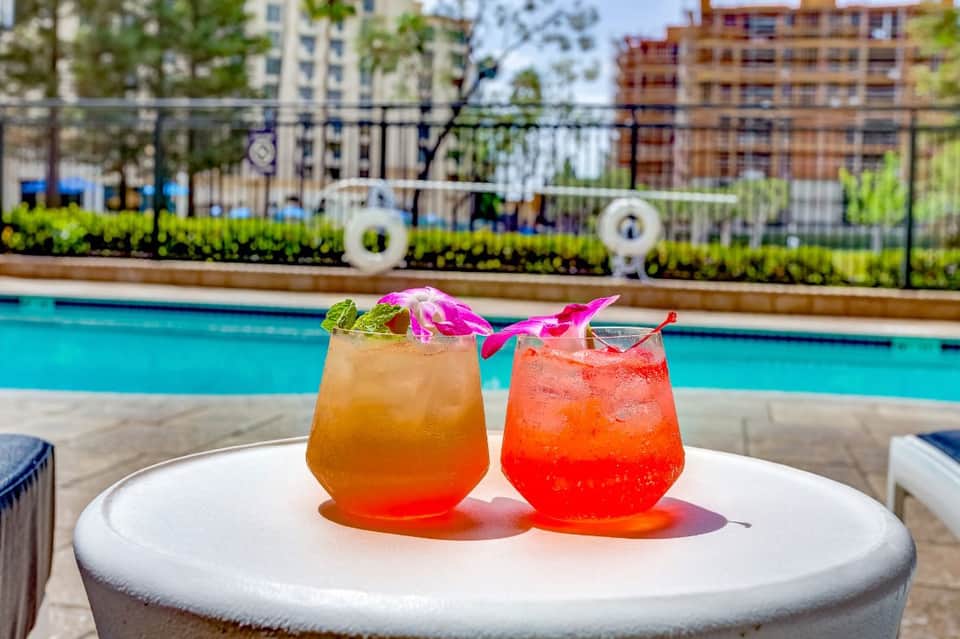 Two colorful cocktails by the pool with tropical flowers and city buildings in the background