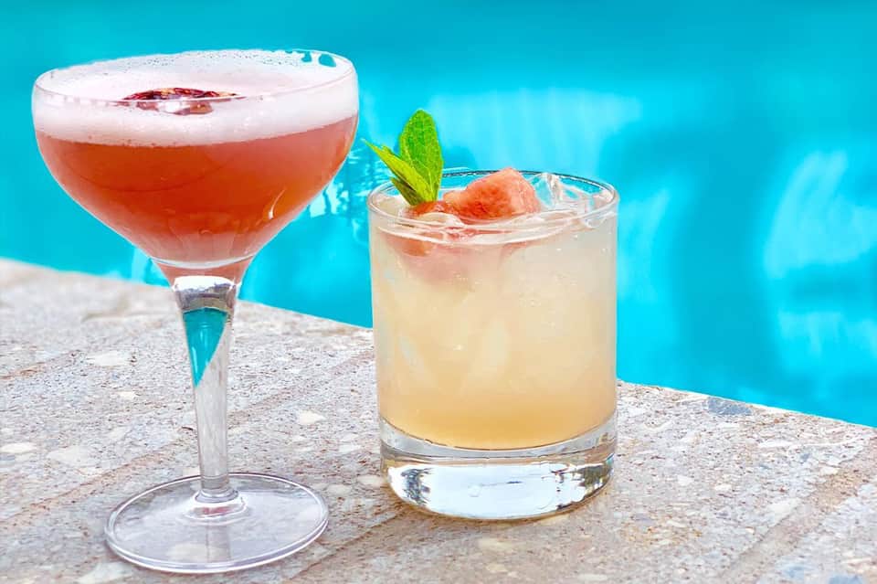 Two colorful cocktails by a turquoise pool: a red drink in a coupe glass and a light cocktail with mint