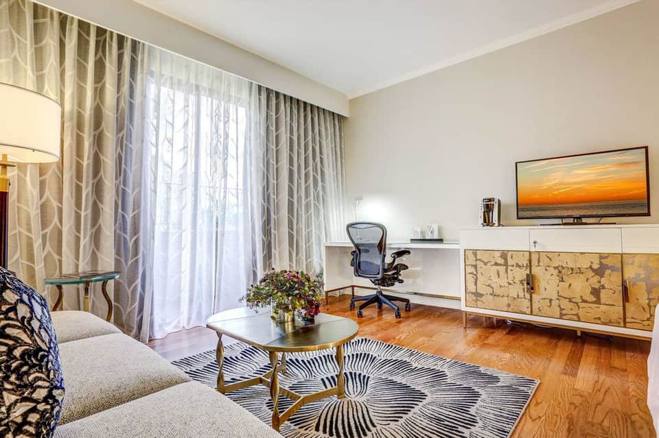 Modern suite with geometric patterned rug, round coffee table, desk with ergonomic chair, and floor-to-ceiling curtains overlooking balcony