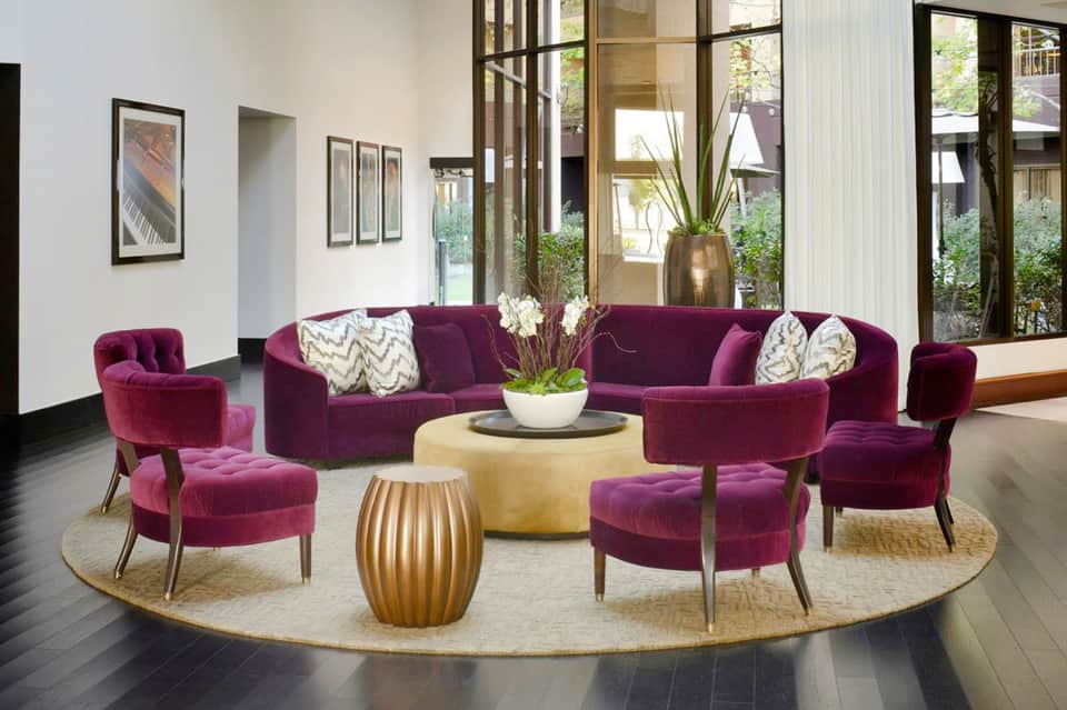 Modern lounge with purple velvet seating, round coffee tables, and large windows overlooking a courtyard garden.