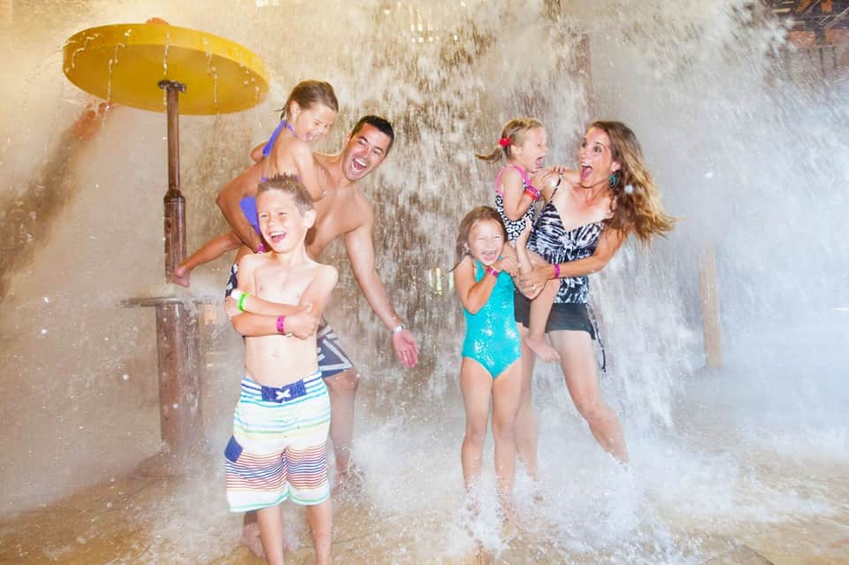 Family enjoying water play under an outdoor shower with yellow umbrella, children and adults splashing together