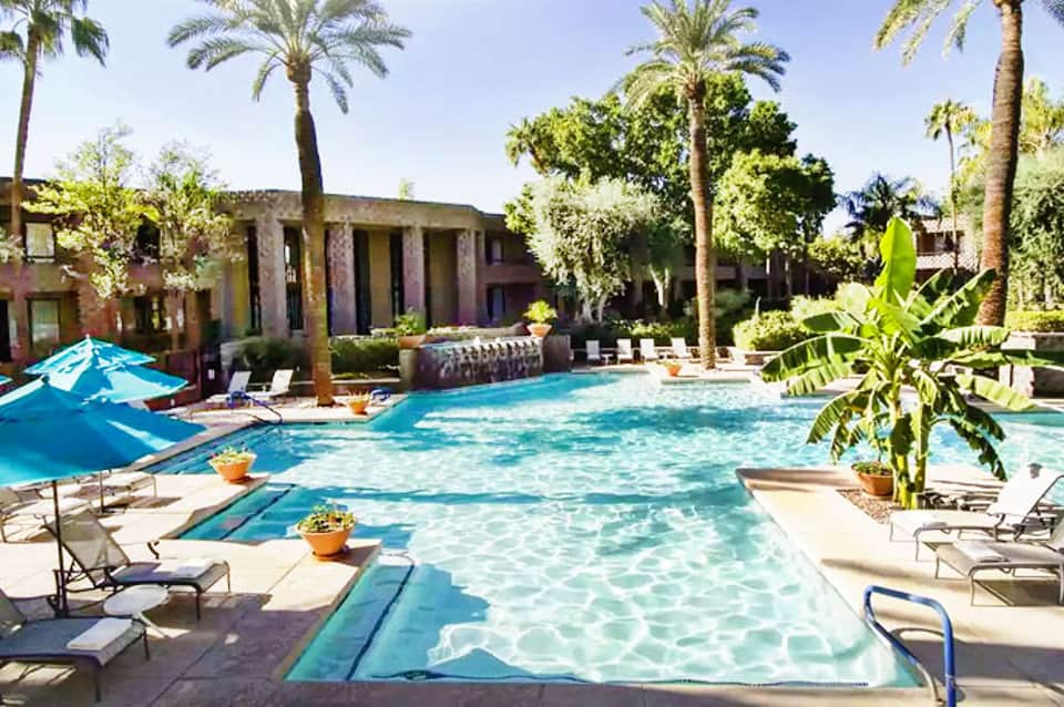 Luxury resort pool surrounded by palm trees with terracotta buildings and lounge chairs in sunny desert setting