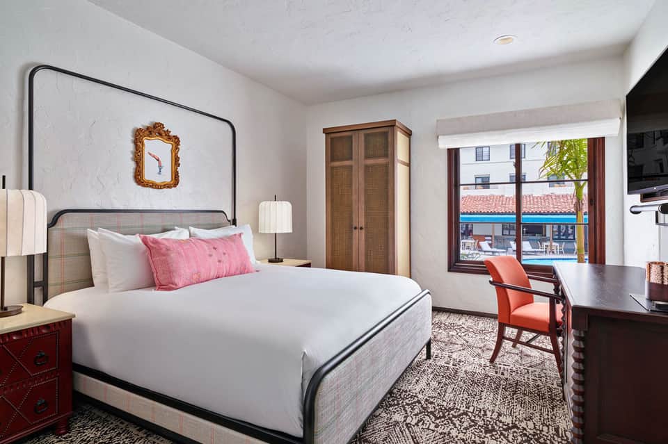 Modern hotel bedroom with white bedding, pink pillows, metal frame bed, and ocean view