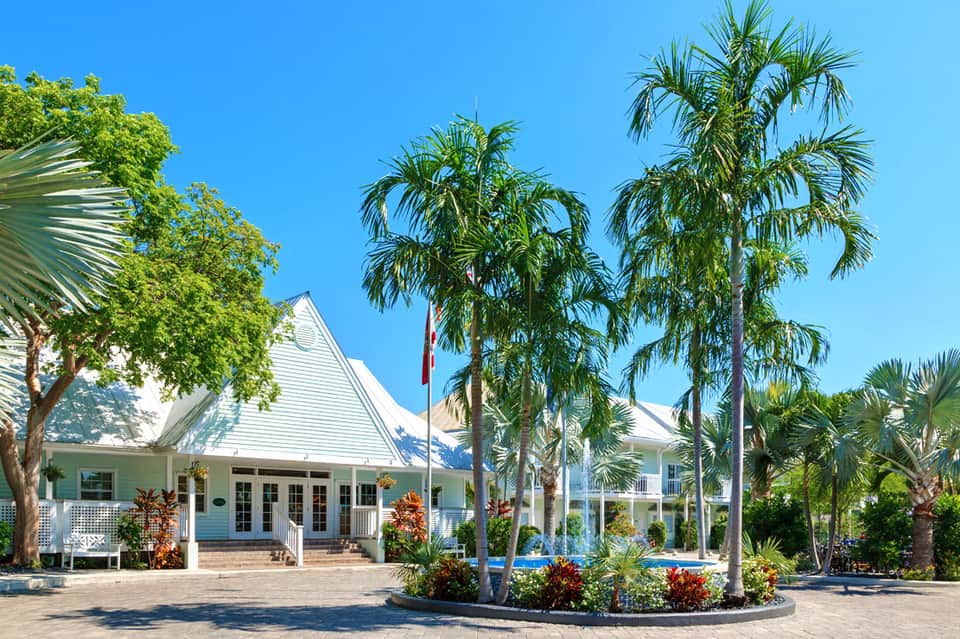 White colonial-style building with wraparound porch, tall palm trees, and manicured landscaping