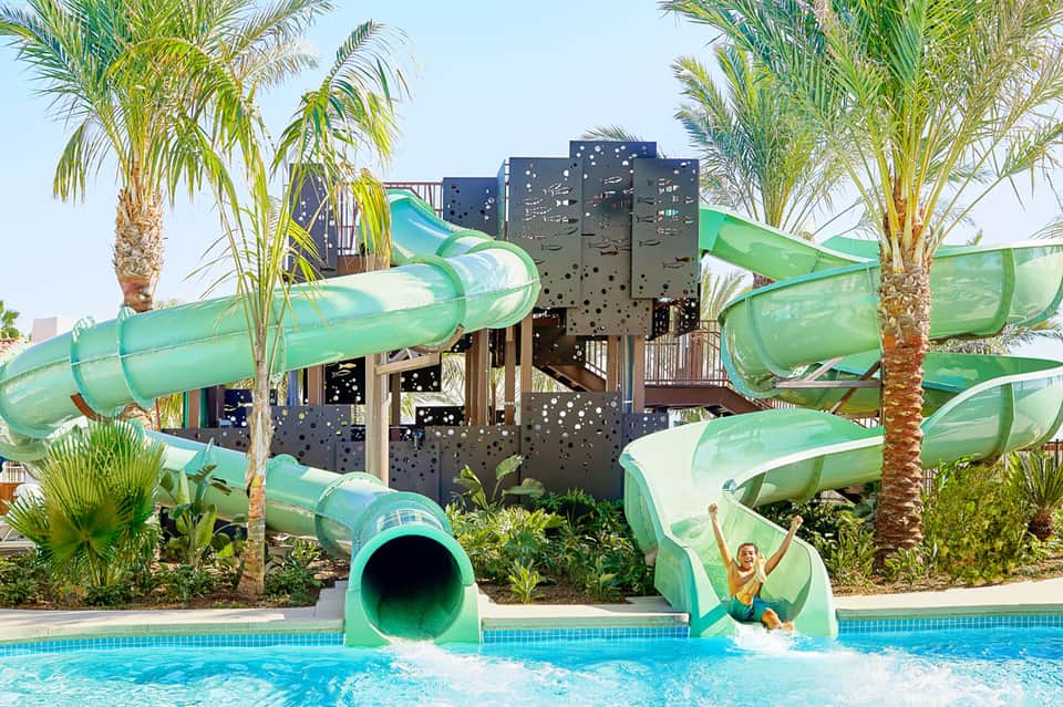 Green water slides at resort pool with palm trees and children playing in turquoise water