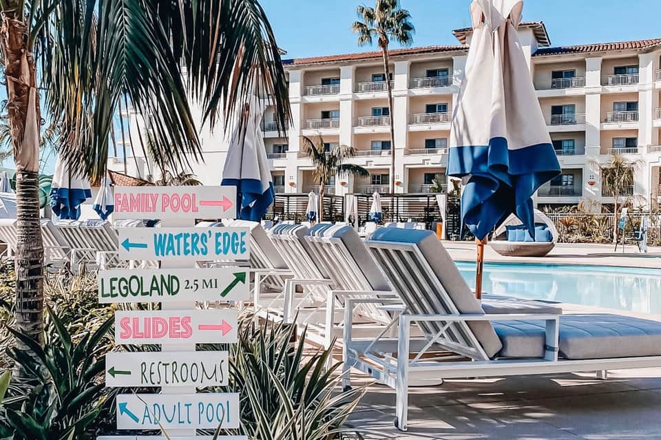 Resort pool with directional signs to family pool, water slide, and adult pool, blue umbrellas, striped lounge chairs, palm trees