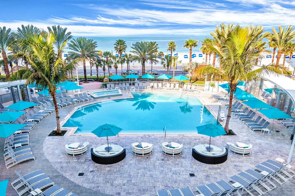 Resort pool deck with turquoise pools, palm trees, lounge chairs, and ocean view beyond