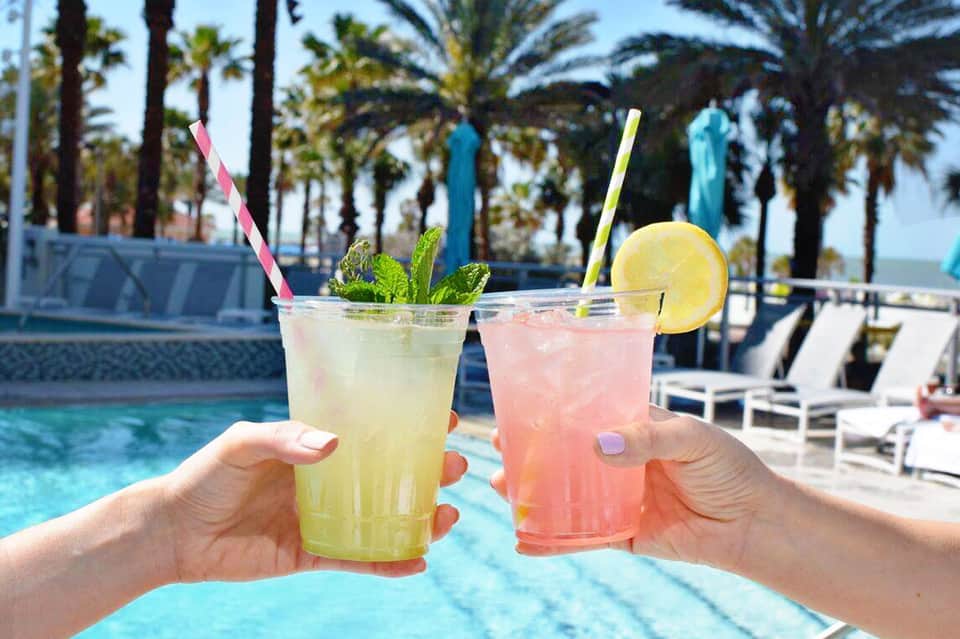 Two colorful cocktails with fresh garnishes by a resort pool and palm trees