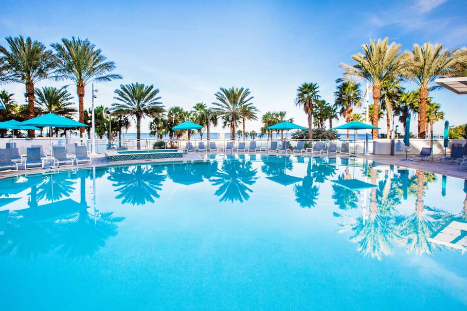 Resort pool with crystal blue water, palm trees, turquoise umbrellas, and beachfront loungers