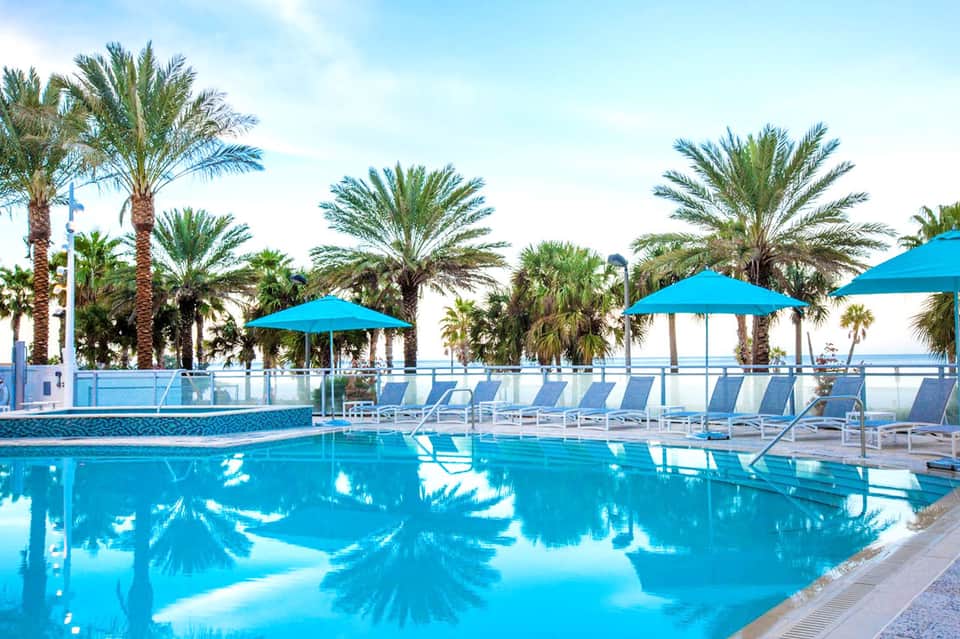 Luxury resort pool with blue umbrellas, palm trees, and ocean view on clear day