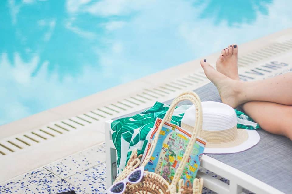 Woman's feet relaxing at pool edge with beach accessories including straw hat, sunglasses, and tote bag