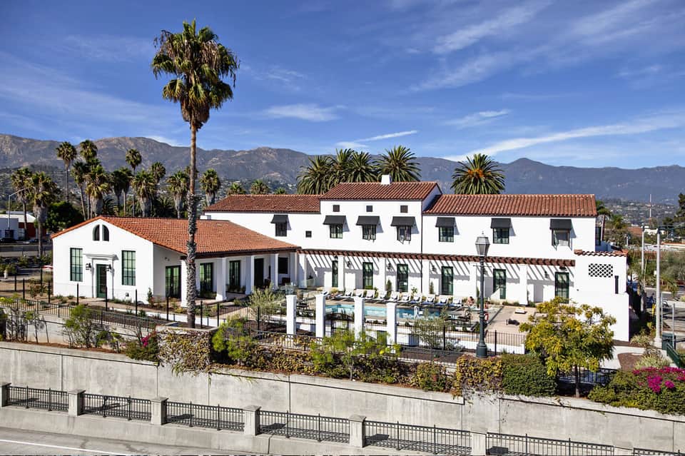 Spanish Colonial-style white hotel with red tile roof, palm trees, and mountains in background