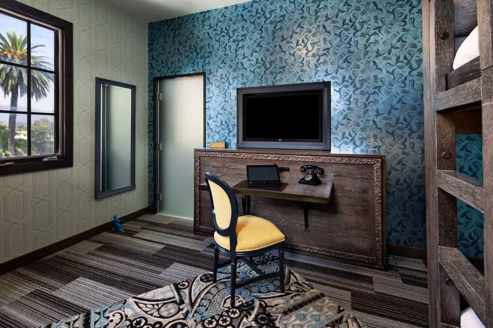 Hotel guest room with blue patterned wallpaper, desk with TV, yellow chair, and window overlooking palm trees