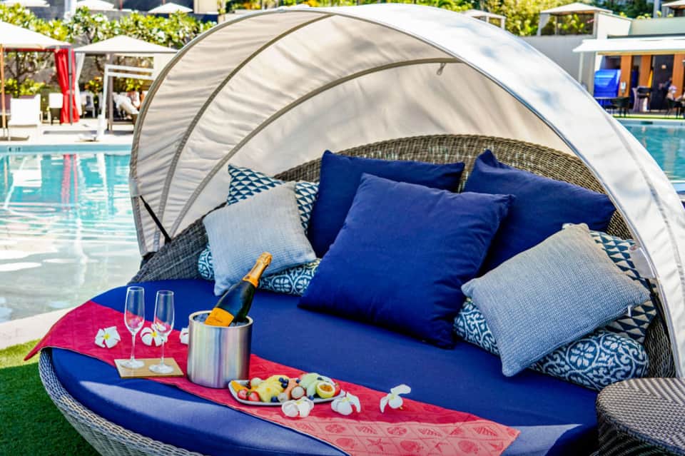 Luxurious poolside cabana bed with canopy, blue cushions, champagne, and fruit platter