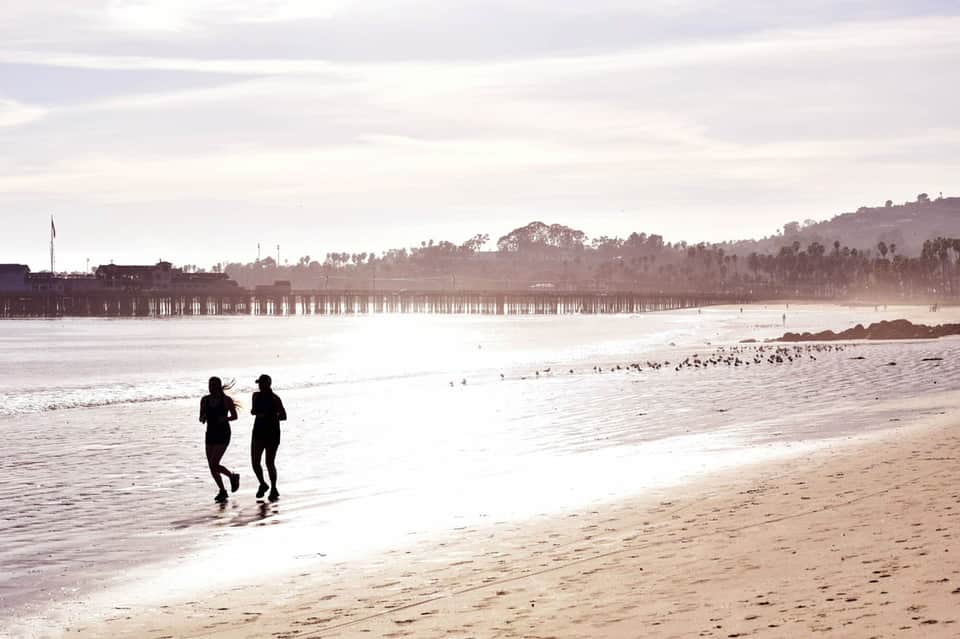 Two runners in wetsuits entering the ocean at a beach with pier and palm trees in background