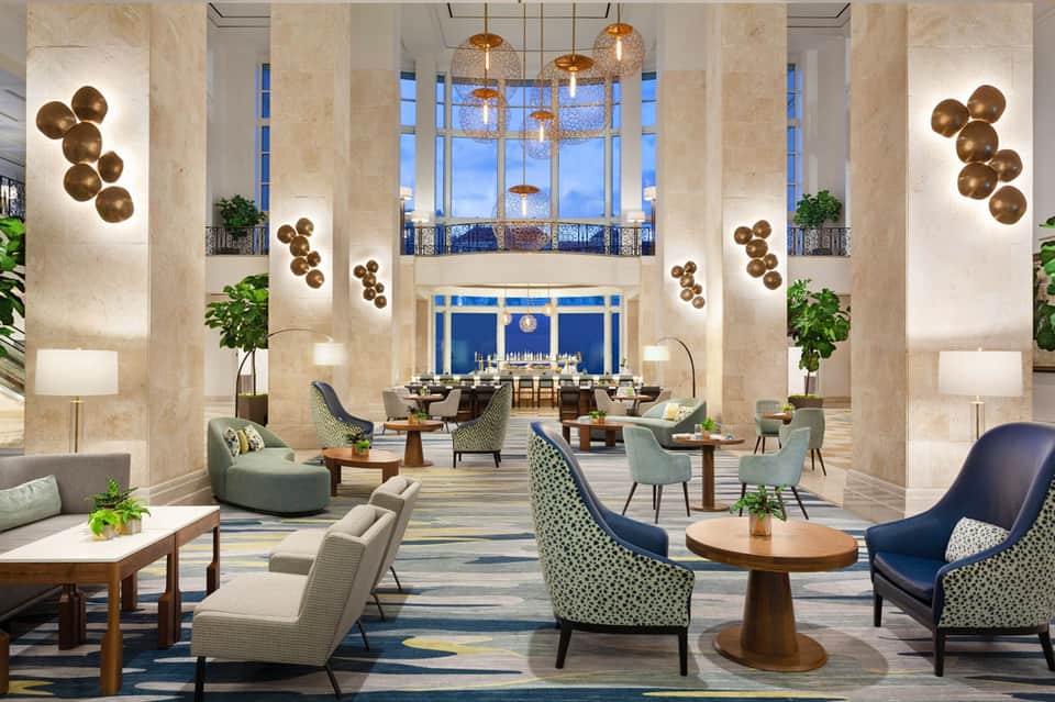Luxury hotel lobby with high ceilings, marble columns, brass pendant lights, and ocean-view windows
