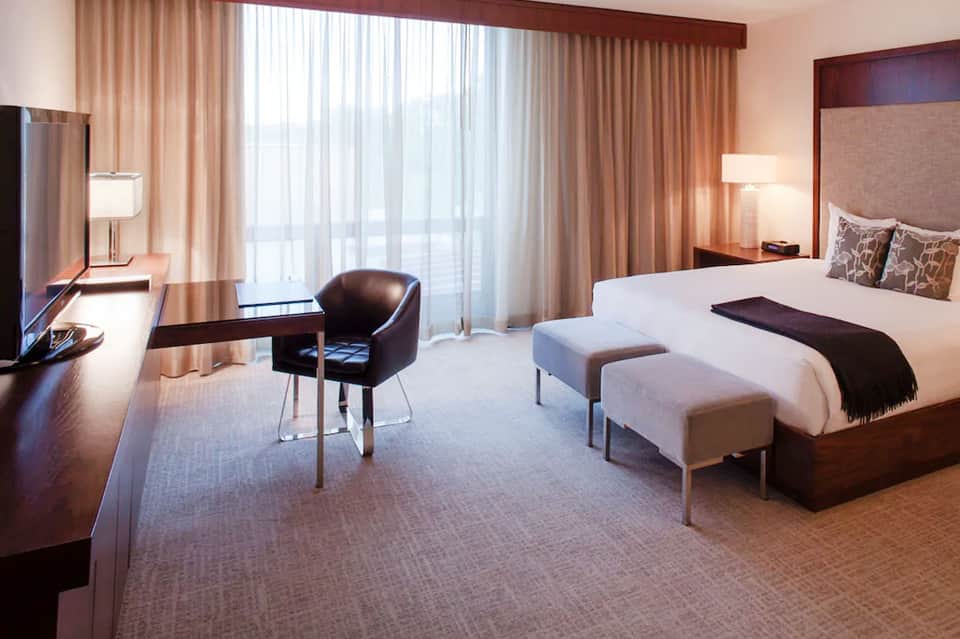 Luxury hotel bedroom with king bed, work desk, dark furnishings, and floor-to-ceiling windows with warm lighting
