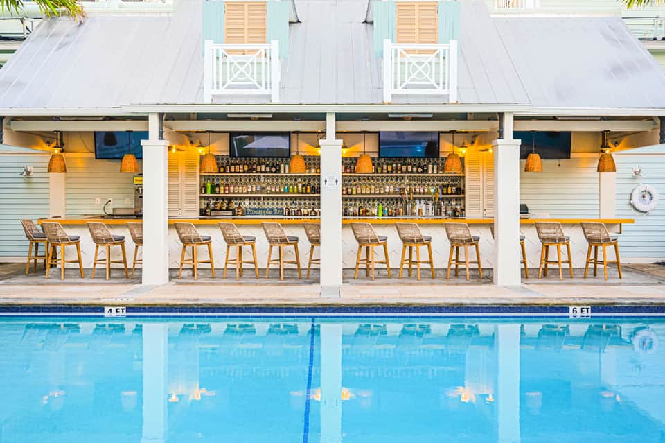 Modern poolside bar with white columns, wooden bar stools, extensive liquor selection, and clear pool water