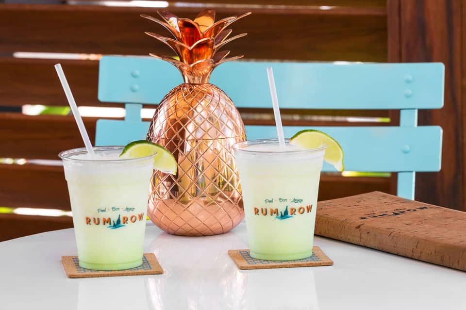 Two tropical cocktails with lime garnish flanking a copper pineapple-shaped drink dispenser on white table