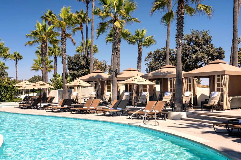 Luxury resort pool with turquoise water, lounge chairs, tan umbrellas, and palm trees under clear blue sky