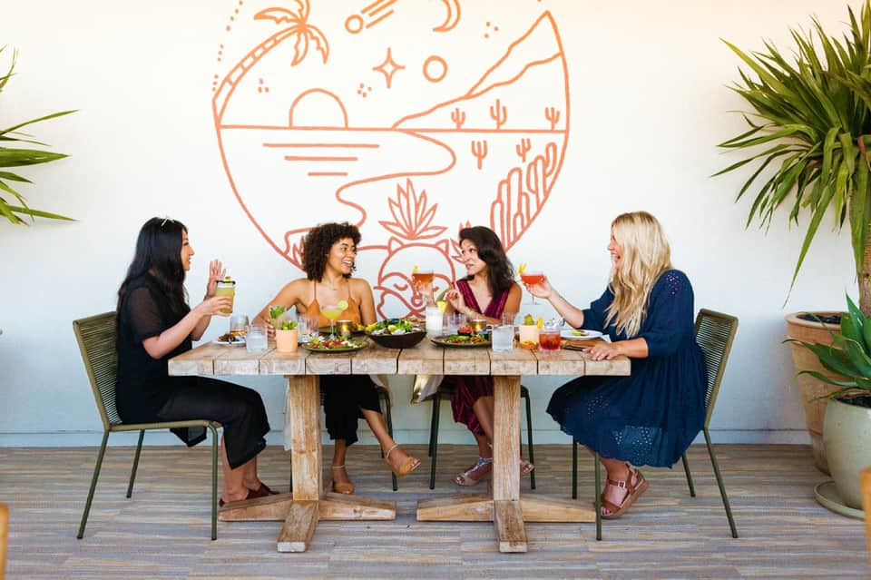 Four women dining together at a wooden table with salads and cocktails, desert mural on wall behind them