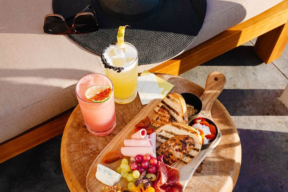 Colorful cocktails and charcuterie board on wooden table by resort lounge chairs