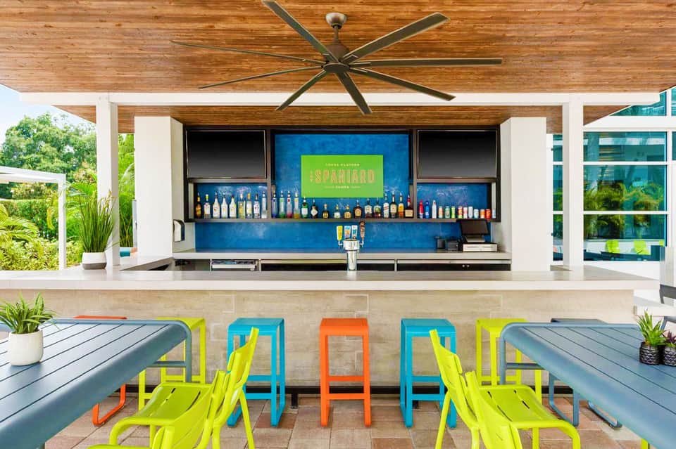 Outdoor bar with blue backdrop, bottle display, and colorful bar stools under wooden pergola with ceiling fan