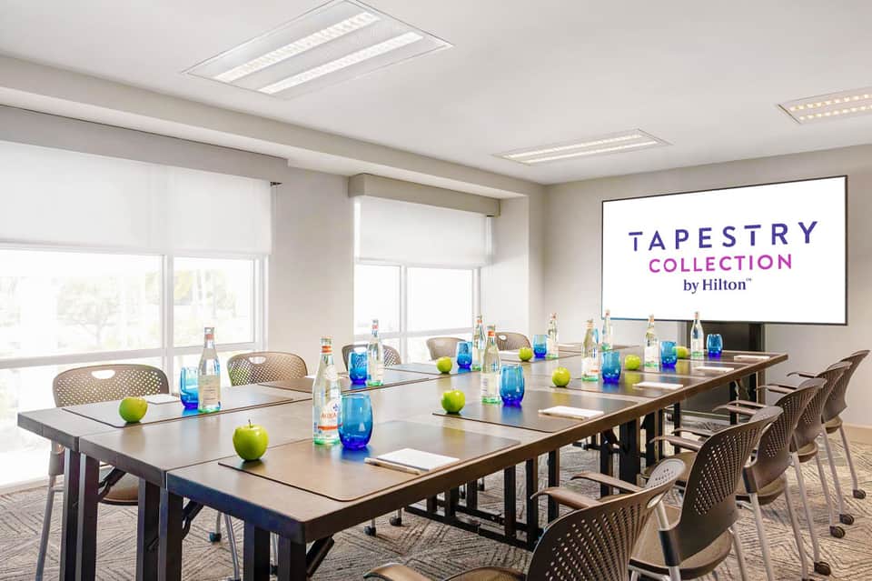 Modern conference room with long table, water bottles, green apples, and Tapestry Collection by Hilton branding on screen