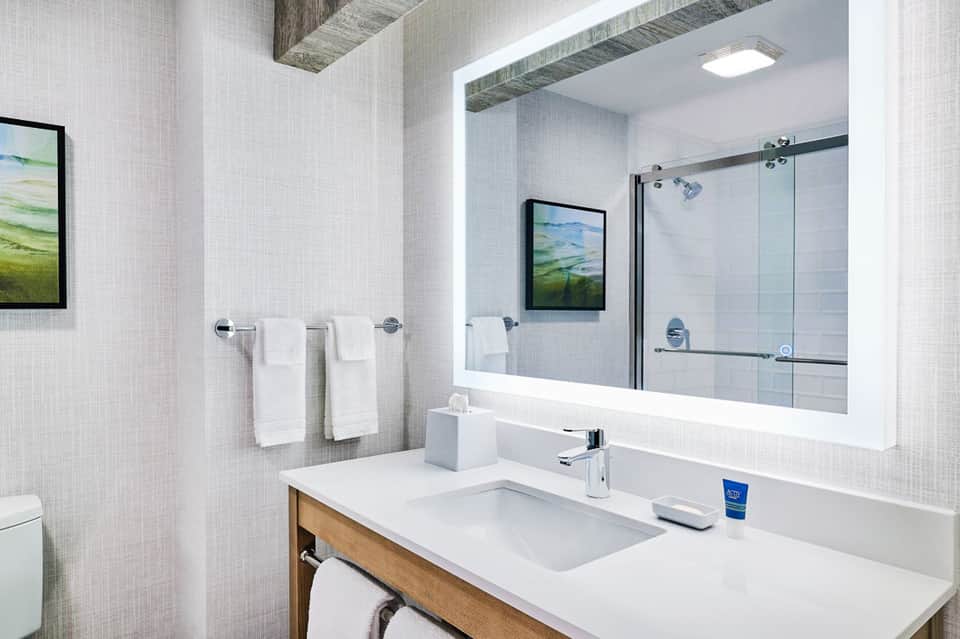 Modern bathroom with illuminated mirror, white vanity, and glass-enclosed shower with landscape artwork