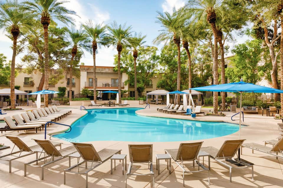Resort pool with loungers, blue umbrellas, palm trees, and Spanish colonial architecture