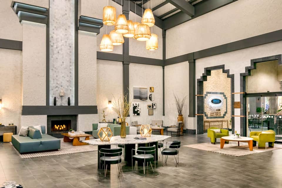 Modern hotel lobby with high ceilings, pendant lights, fireplace, and contemporary furniture in green and yellow tones