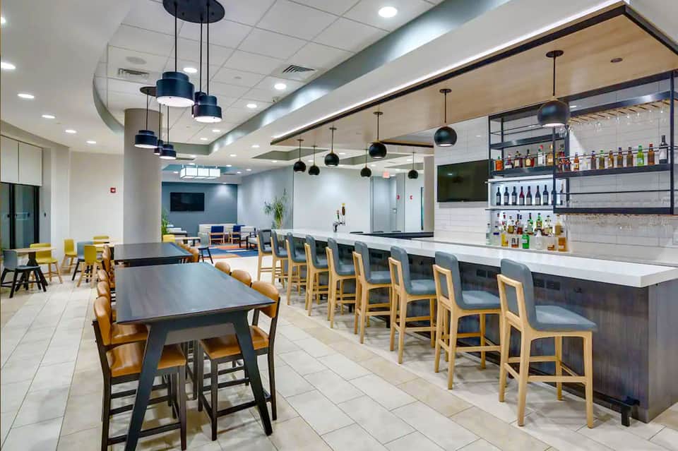 Modern hotel bar and lounge with high ceilings, pendant lighting, wood chairs, and bottle display shelves