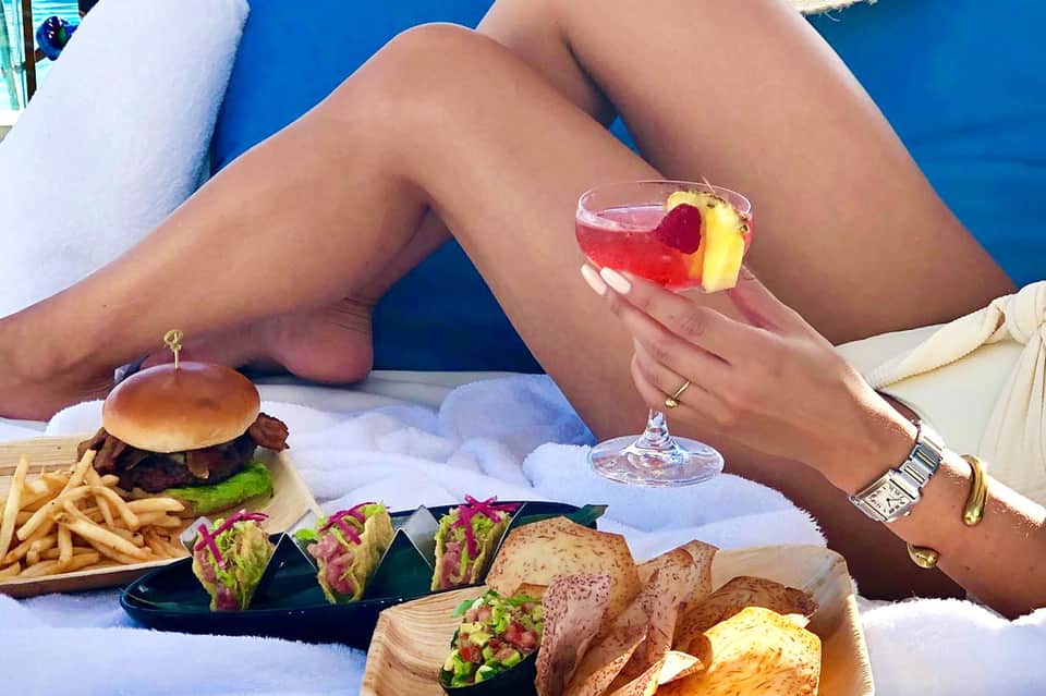 Woman relaxing by pool holding colorful cocktail with gourmet food spread nearby