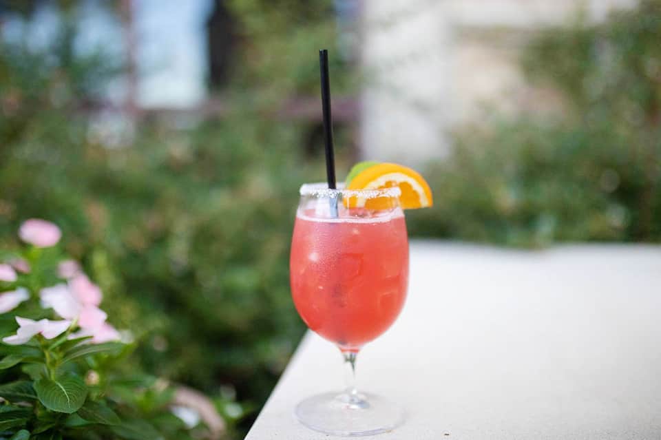 Coral-colored cocktail with orange slice garnish on outdoor table with greenery backdrop
