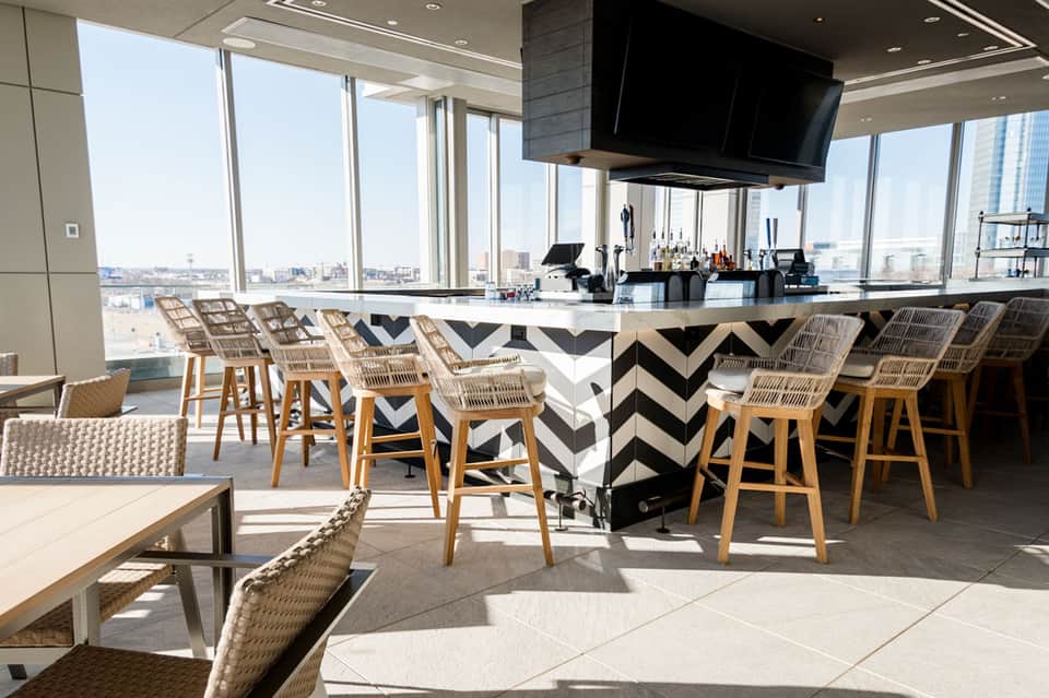 High-rise rooftop bar with chevron-patterned counter, woven bar stools, and expansive city skyline views