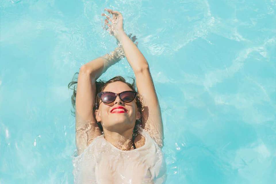 Woman floating in a turquoise pool wearing sunglasses and a white shirt