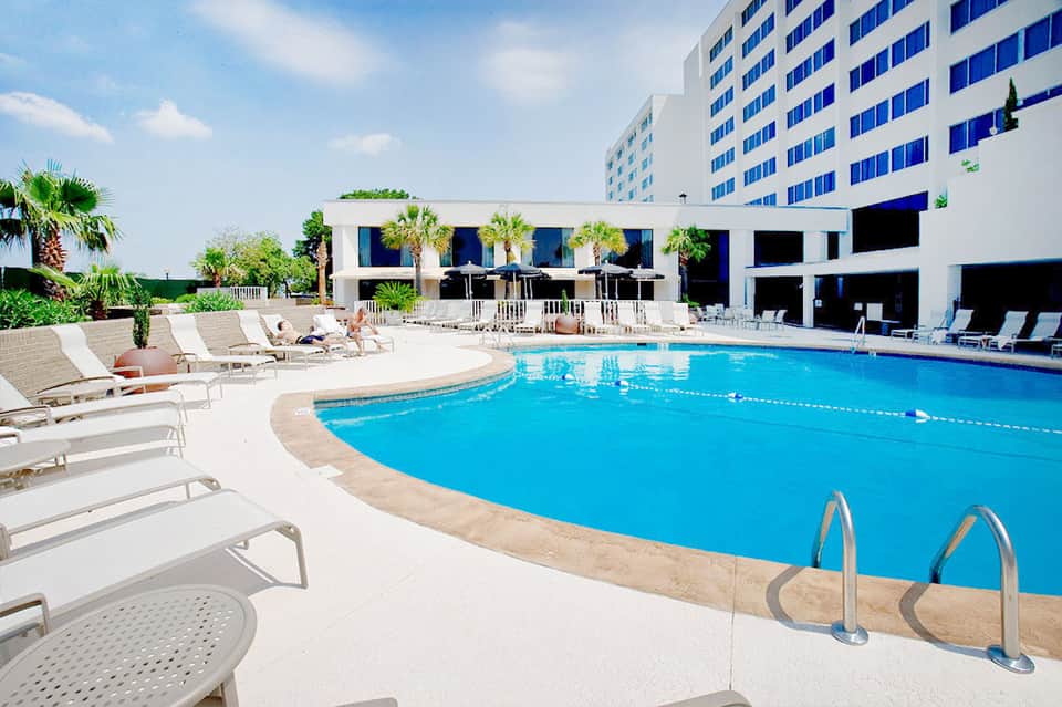 Curved resort pool with lane markers, lounge chairs, and multi-story hotel building under blue sky