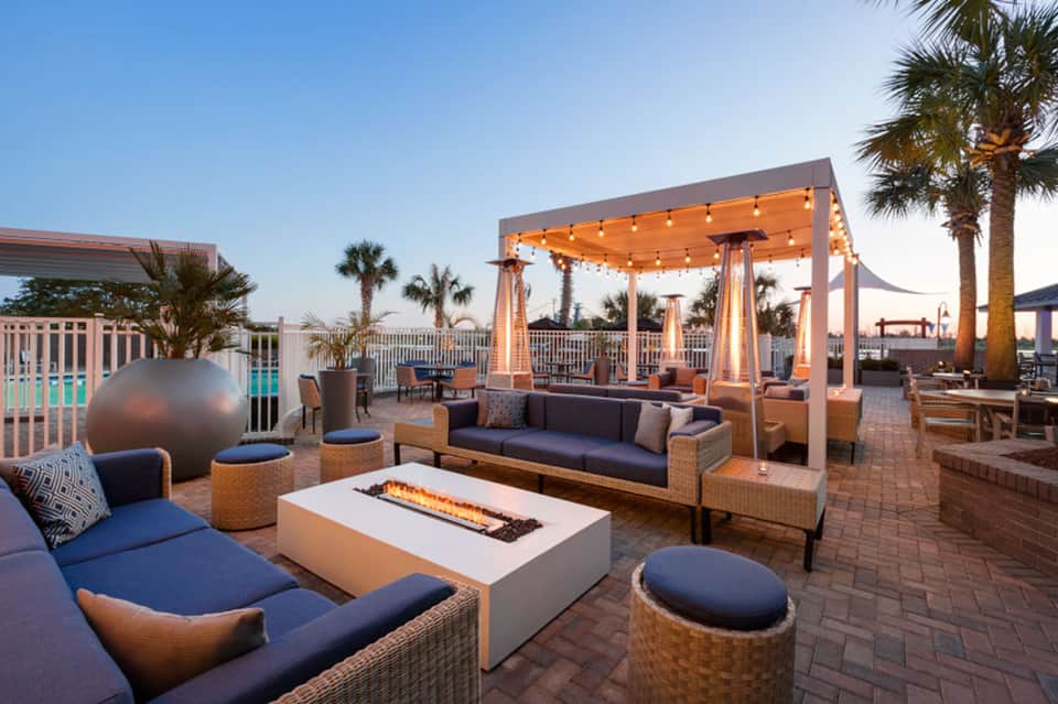 Rooftop lounge at dusk with fire pit, woven furniture, wooden pergola, and string lights overlooking palms