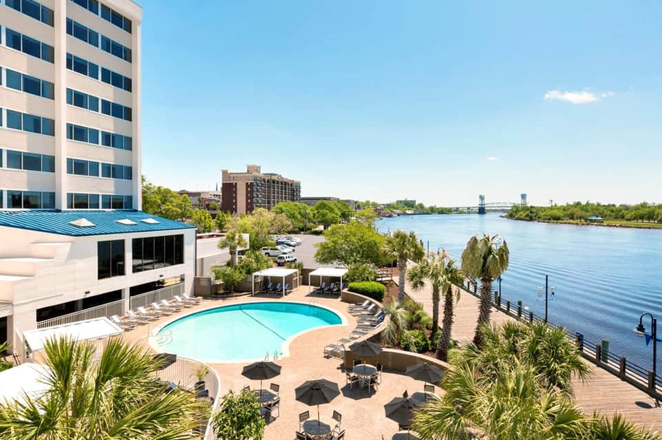 Waterfront hotel with pool, cabanas, palm trees, and river views under clear blue sky