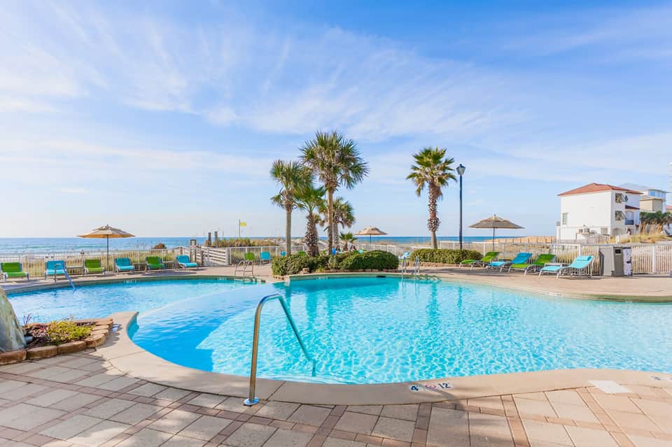 Beachfront resort pool with palm trees, umbrellas, and ocean view under clear blue sky
