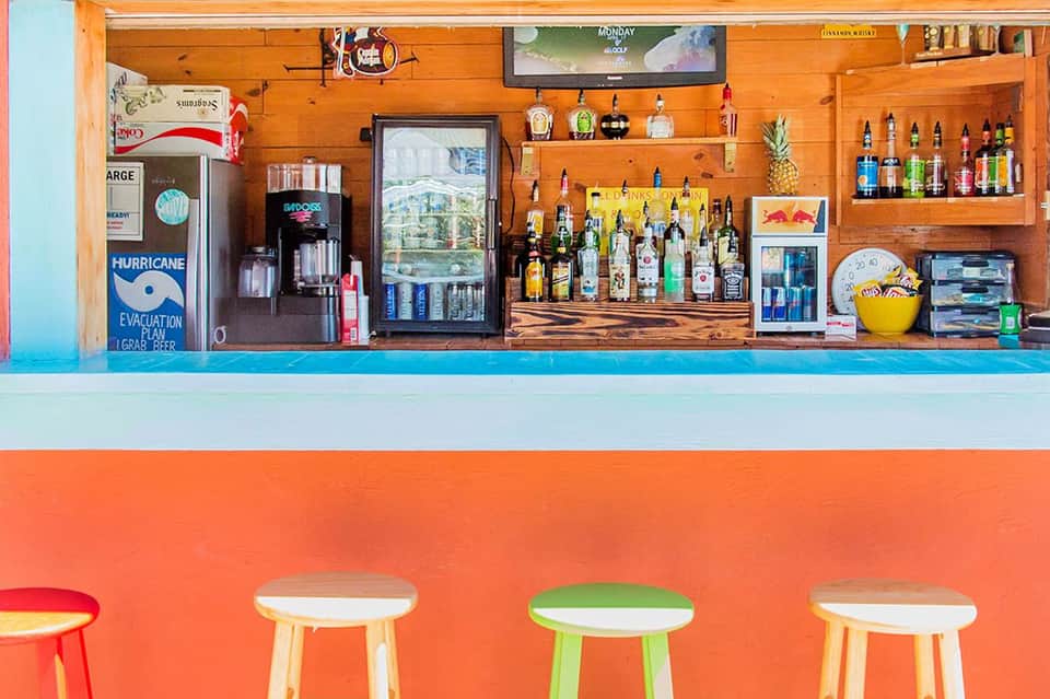 Colorful beach bar with orange exterior, wooden interior stocked with bottles, and rainbow stools
