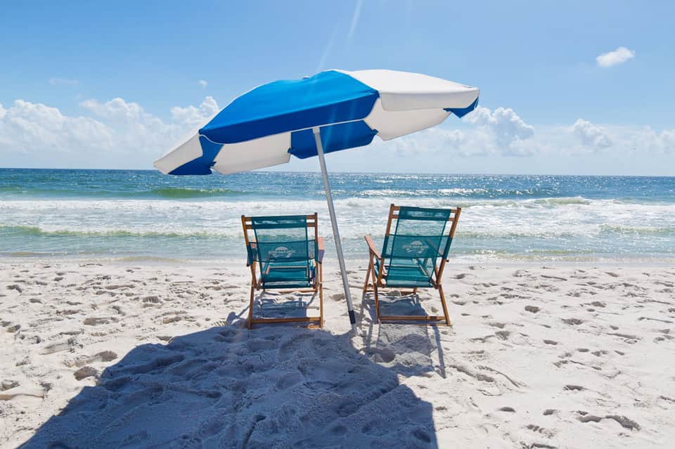 Two teal beach chairs under a blue and white striped umbrella on sandy beach with ocean waves