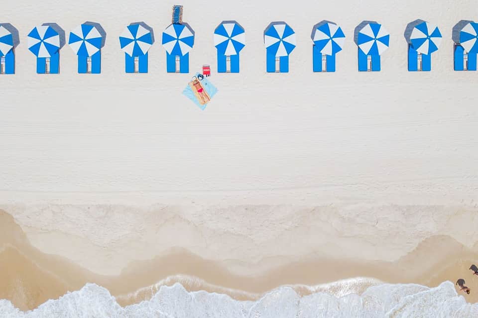 Aerial view of a lone person on a beach towel surrounded by blue and white beach umbrellas
