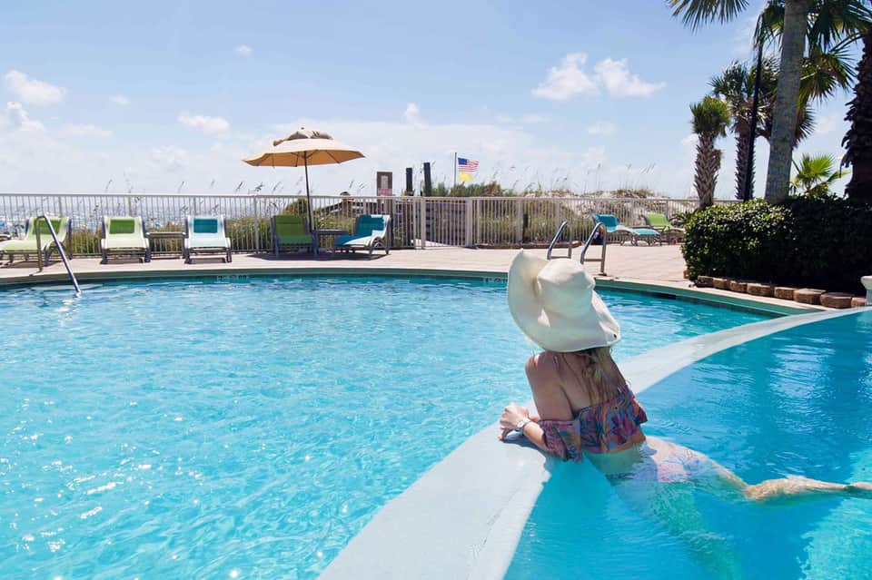 Guest swimming in crystal-clear pool with ocean view, palm trees, and colorful lounge chairs