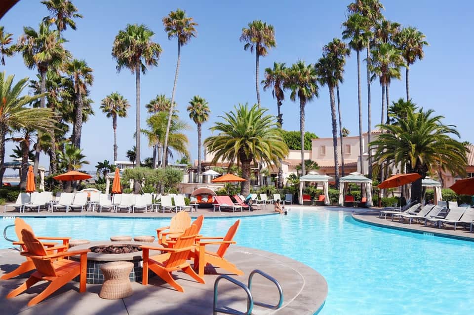 Resort pool area with palm trees, orange Adirondack chairs, fire pit, and loungers under umbrellas