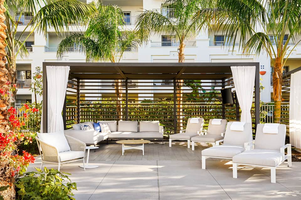 Luxury resort patio with covered pergola, gray sectional sofa, white lounge chairs, and palm trees