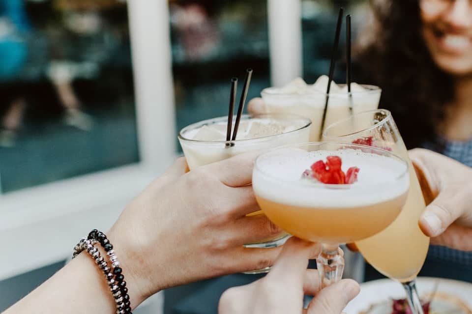 Hands holding craft cocktails with foam and garnish overlooking an indoor pool area