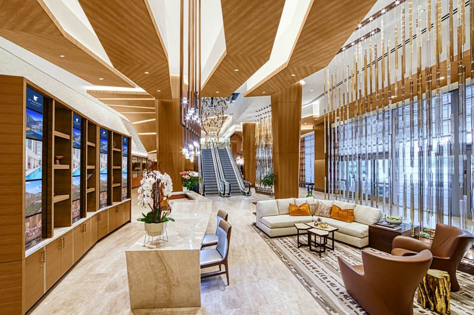 Luxurious modern hotel lobby with wooden ceiling panels, marble flooring, escalators, and contemporary seating areas