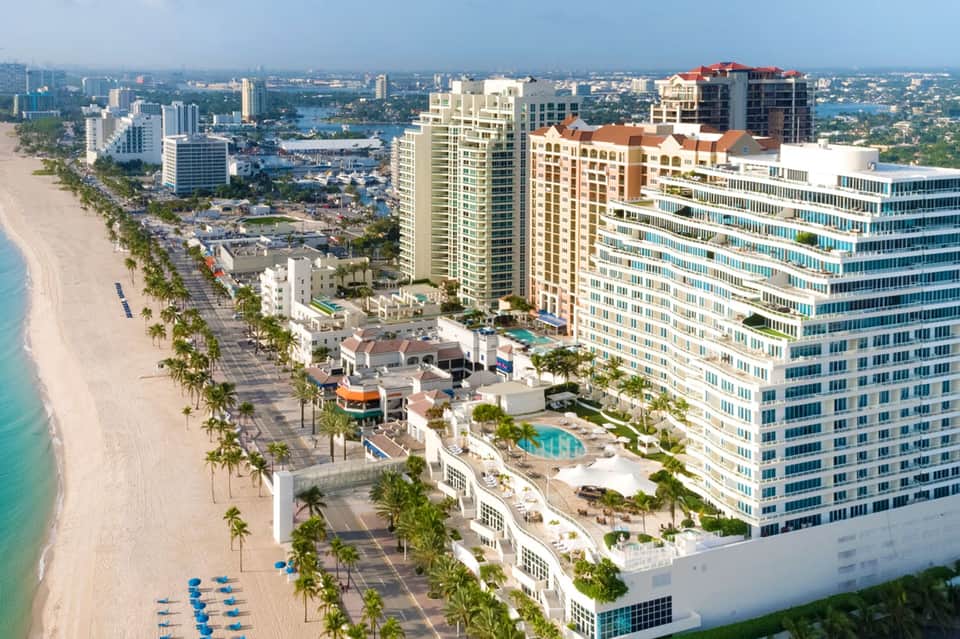 Beachfront resort aerial view with high-rise towers, sandy beach, palm trees, and distant waterway