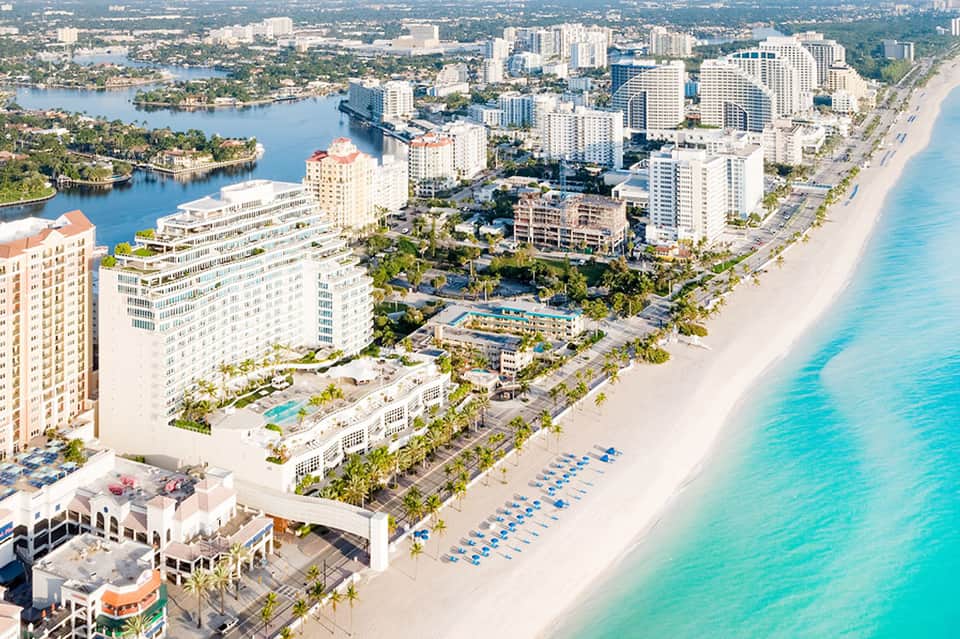 Beachfront resort aerial view showing turquoise ocean, sandy beach, high-rise hotels, and palm trees